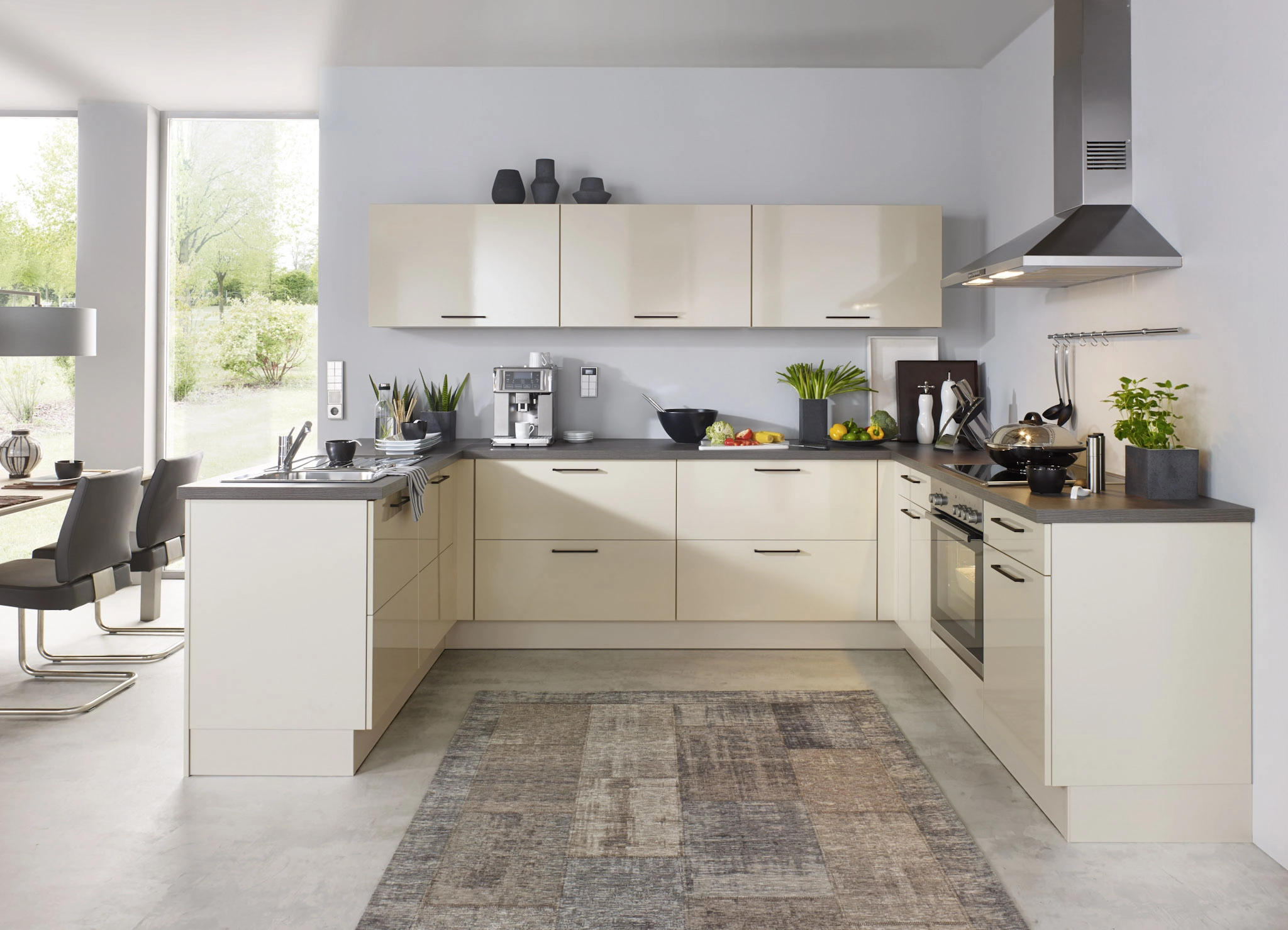Cuisine moderne en U beige brillant avec plan de travail gris foncé, hotte design en inox et grand tapis décoratif.