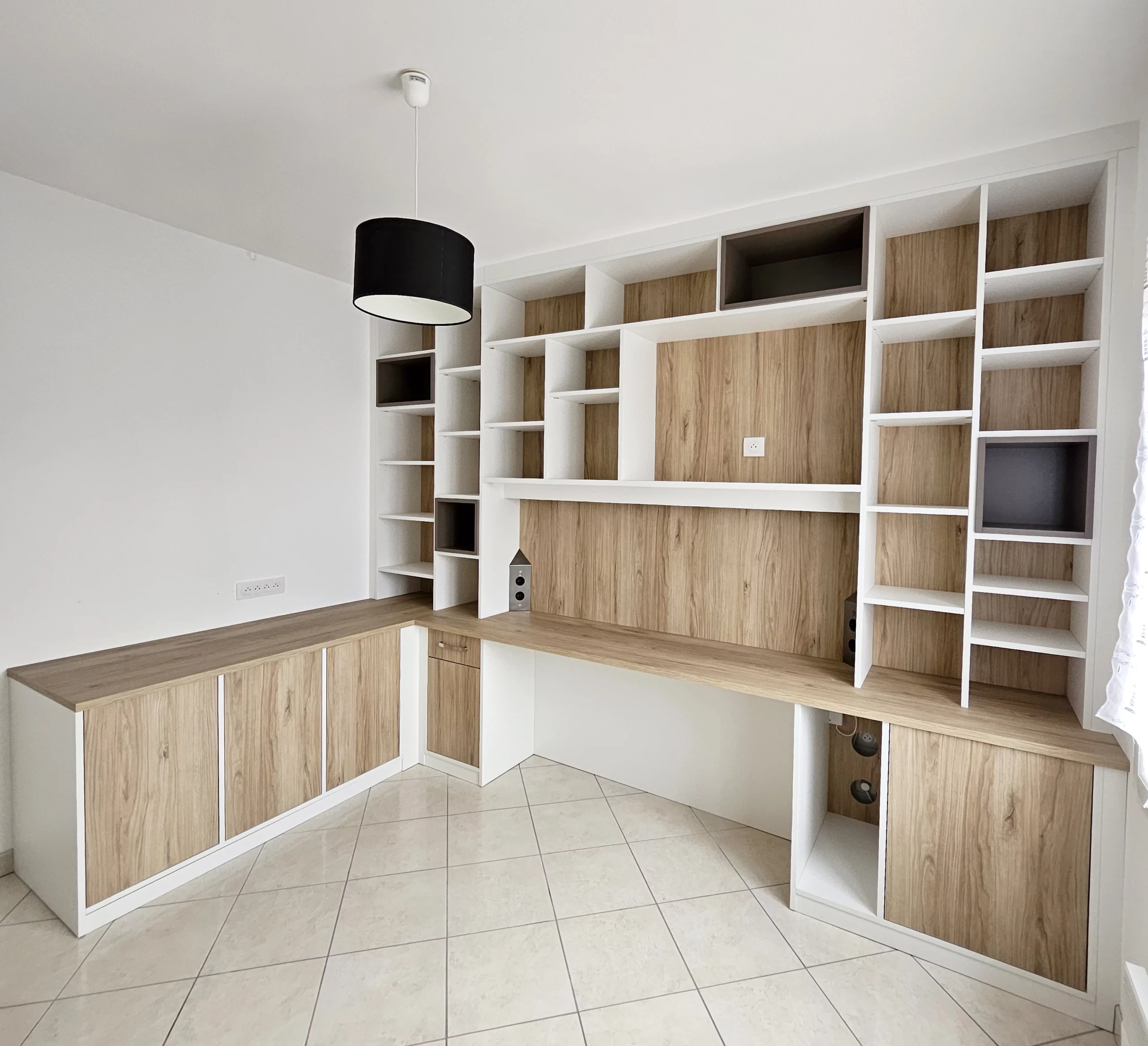 Vue d'angle d'un grand bureau d'angle sur mesure intégrant un plan de travail en bois clair, des meubles bas blancs et une vaste bibliothèque murale toute hauteur.