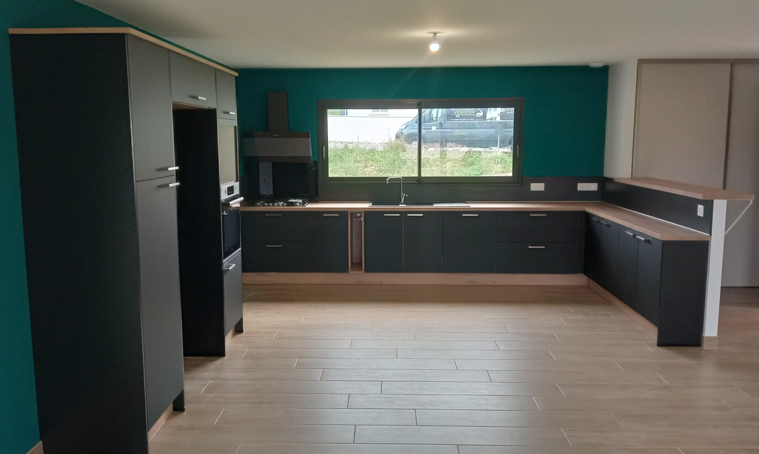 Cuisine moderne en U bleu canard et bois : Vue d'ensemble d'une cuisine équipée avec des façades noir mat, un plan de travail en bois clair et un mur d'accent bleu canard, incluant un coin repas bar.