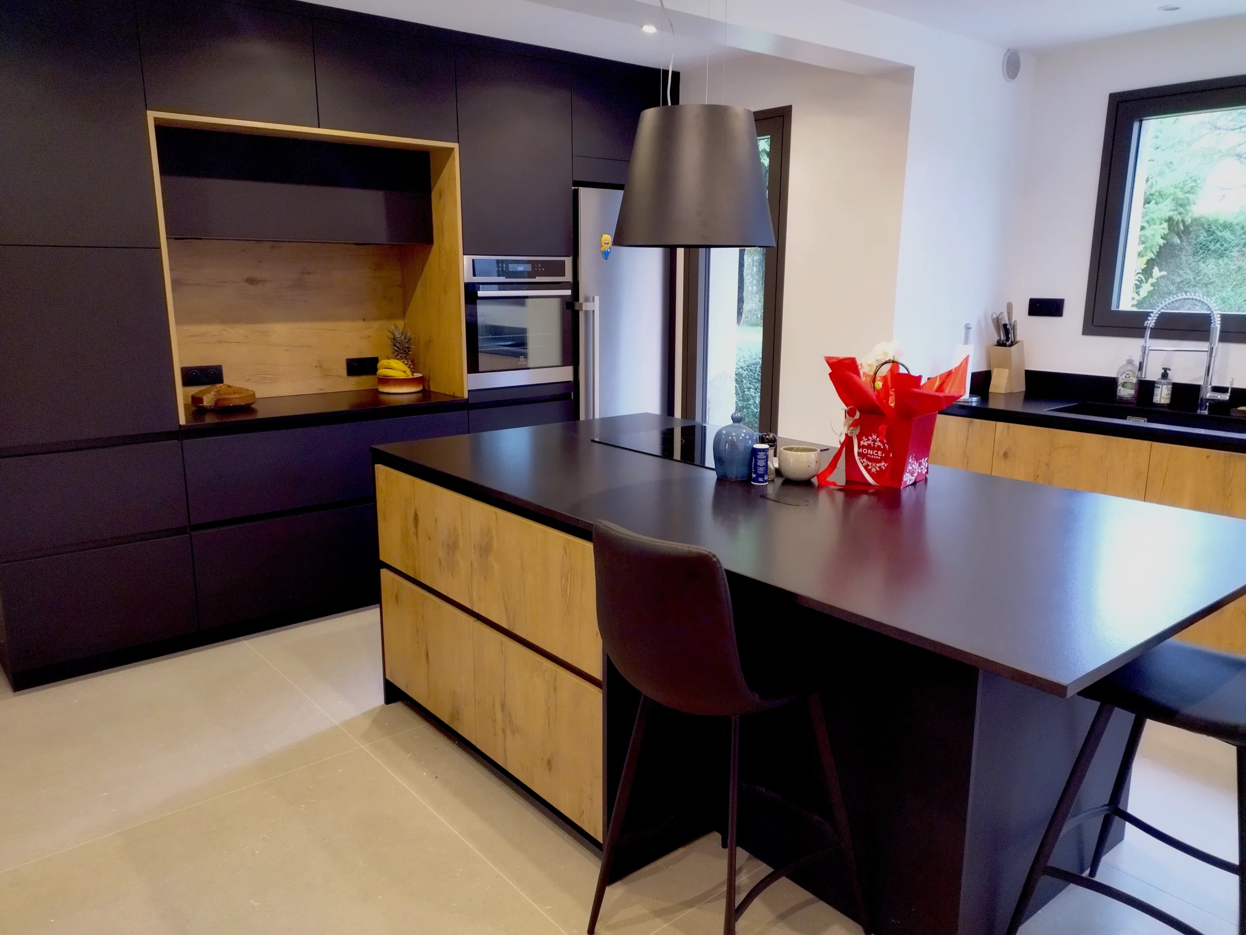 Zoom sur îlot de cuisine avec plaque intégrée : Vue rapprochée d'un îlot central noir mat équipé d'une plaque de cuisson, de tiroirs de rangement et d'une étagère latérale en bois.