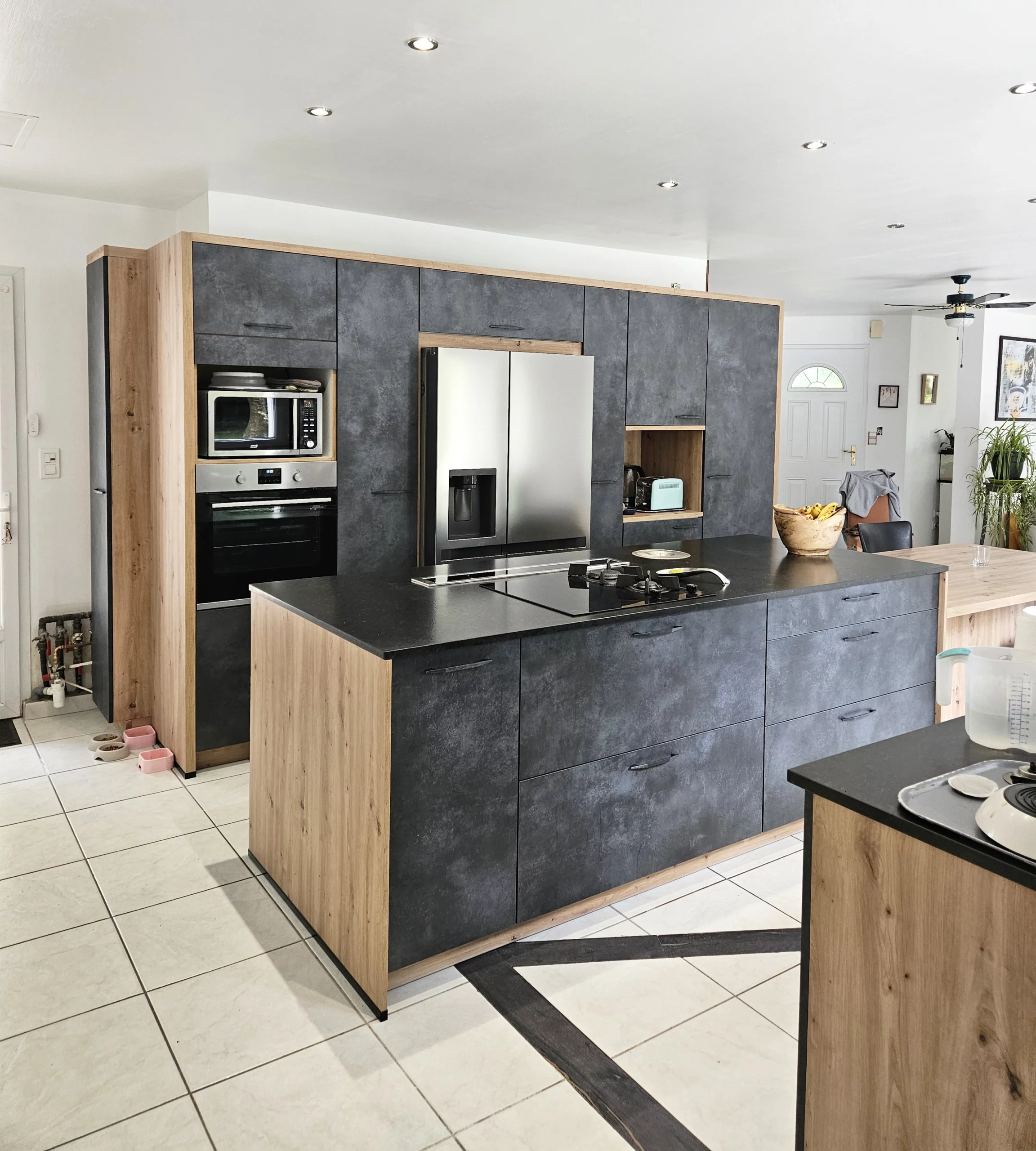 Cuisine moderne avec îlot central en pierre sombre : Vue d'ensemble d'une cuisine équipée avec de grandes colonnes de rangement en bois et gris anthracite, incluant un îlot central massif et un réfrigérateur américain.