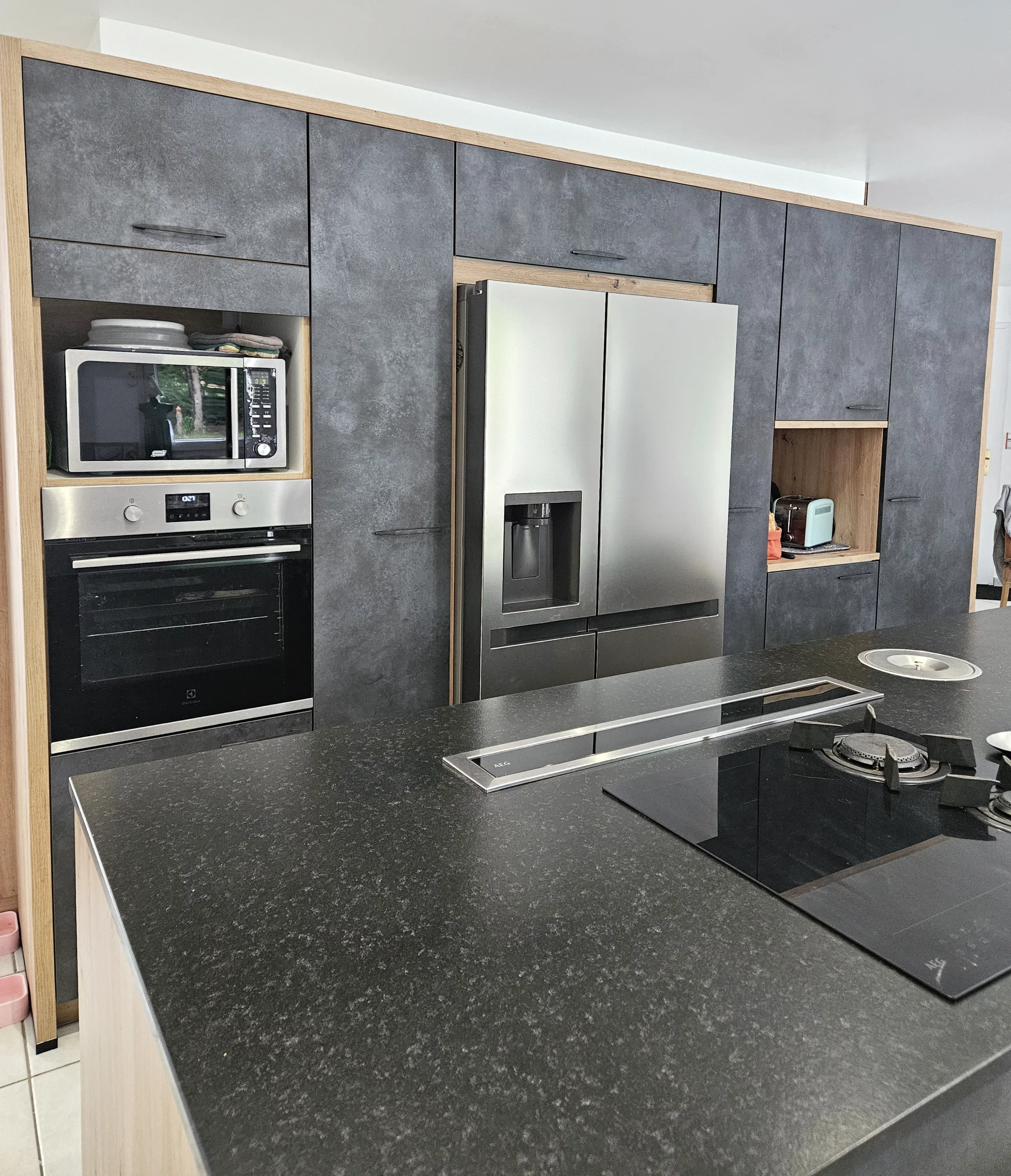 Plan de travail avec plaque de cuisson et hotte intégrée : Zoom sur l'îlot central de cuisine noir mat avec une plaque de cuisson moderne et un système d'aspiration intégré, devant un mur de rangements gris.