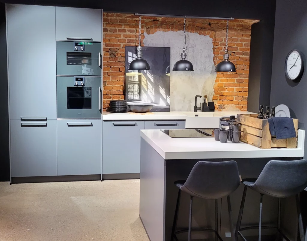 Cuisine moderne bleu gris installée devant un authentique mur de briques rouges avec luminaires industriels.