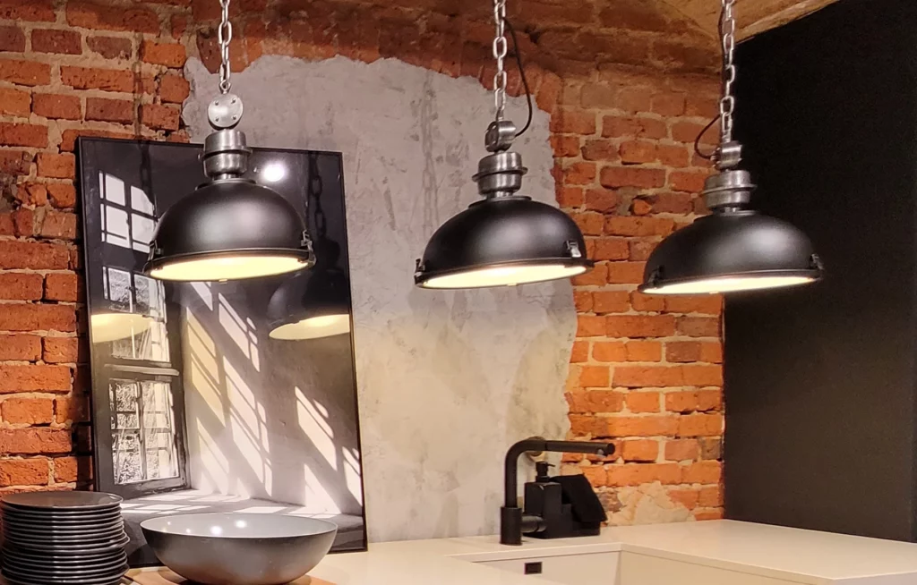 Zoom sur le plan de travail d'une cuisine industrielle avec évier noir, suspensions métalliques et mur de briques.