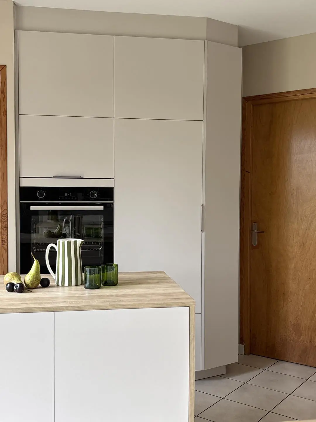 Colonnes de rangement toute hauteur avec façades claires et four encastré, associées à un îlot en bois miel pour un équilibre visuel apaisant.