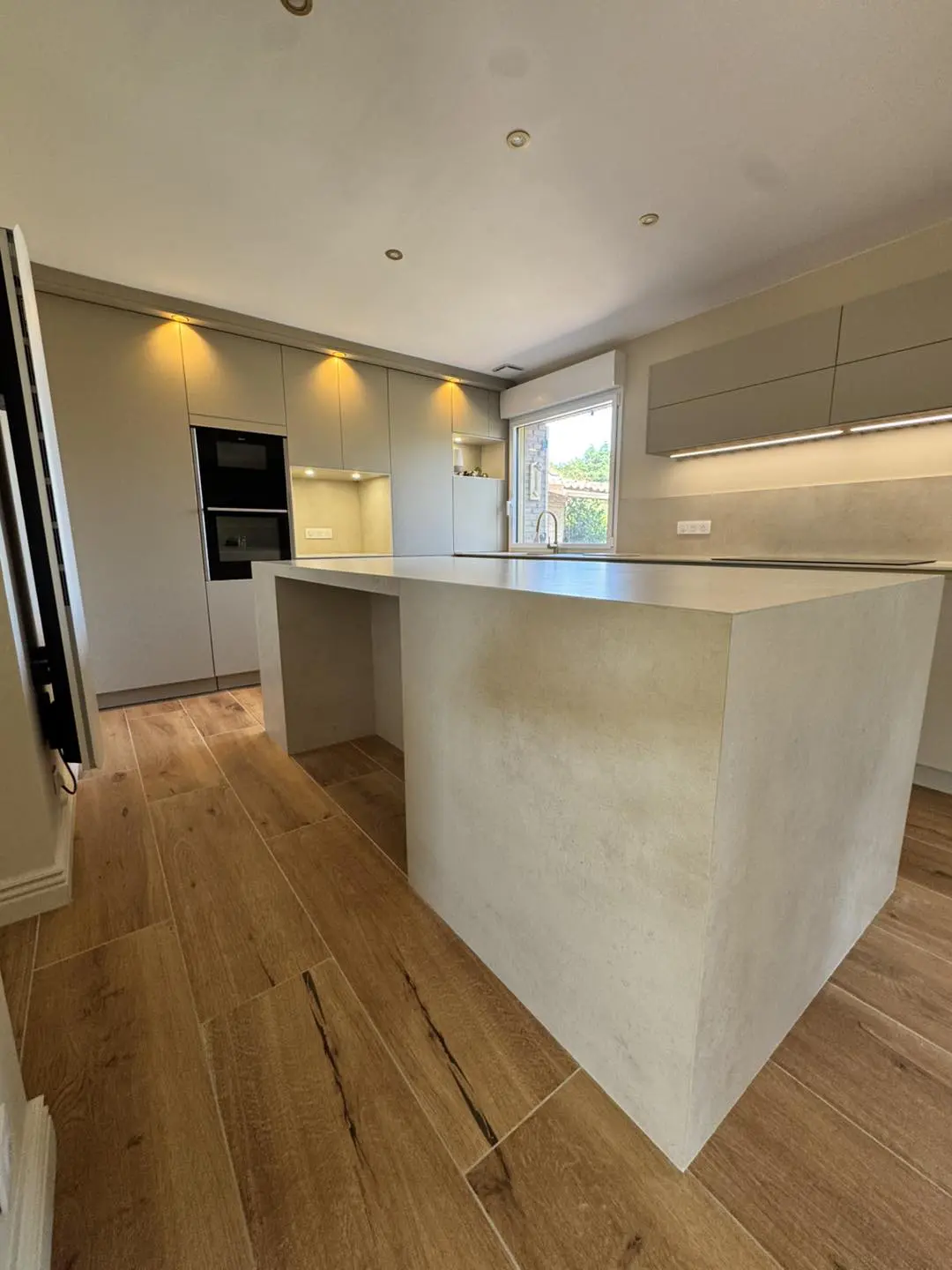 Aménagement de cuisine contemporaine mêlant matières brutes et teintes apaisantes, avec îlot en céramique et éclairage sous meubles hauts.