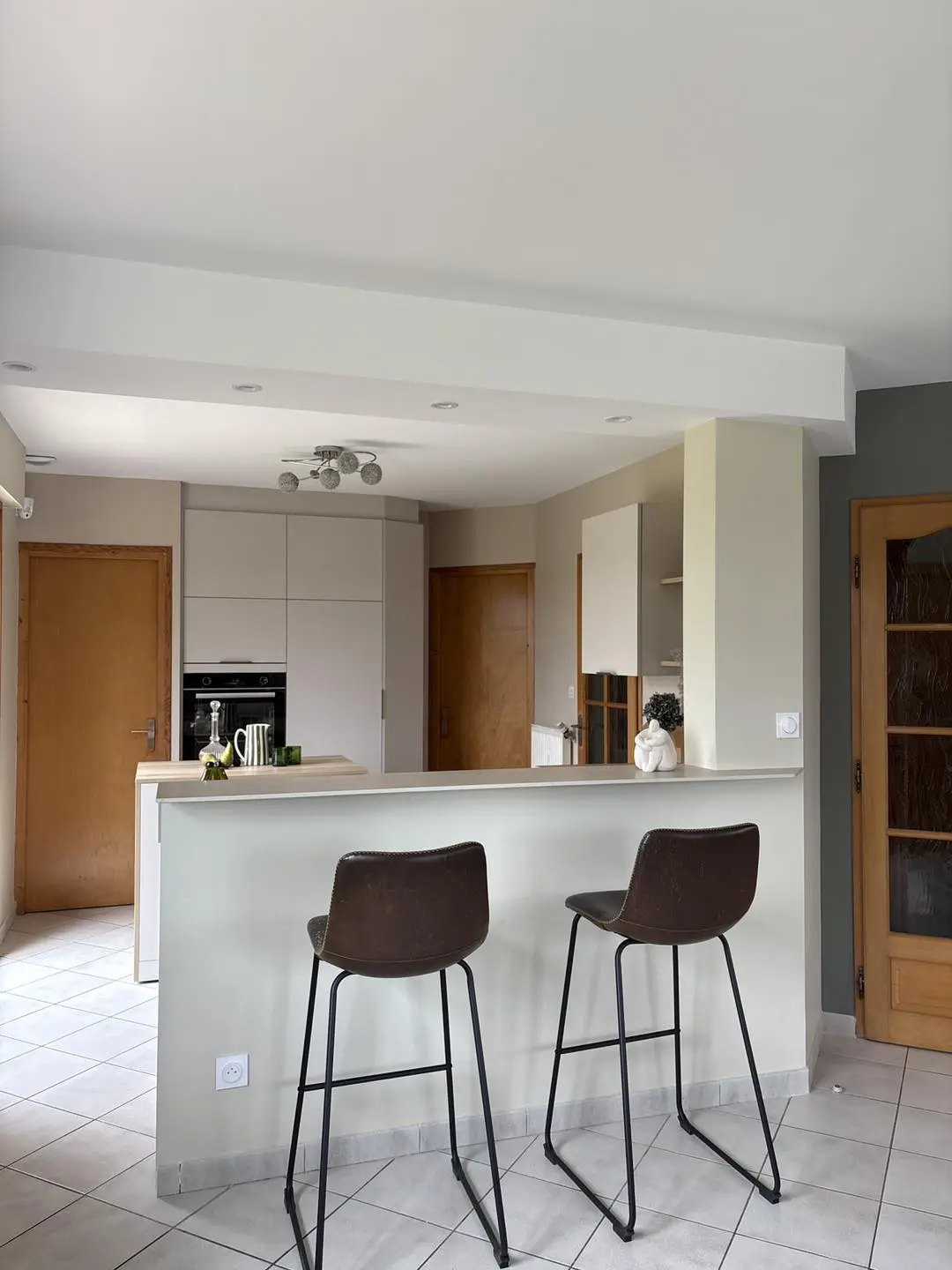 Transformation d'une cuisine fermée en un espace ouvert et lumineux avec bar dînatoire, tabourets en cuir et plan de travail en bois chaleureux, réalisée par Maximilien Lust.