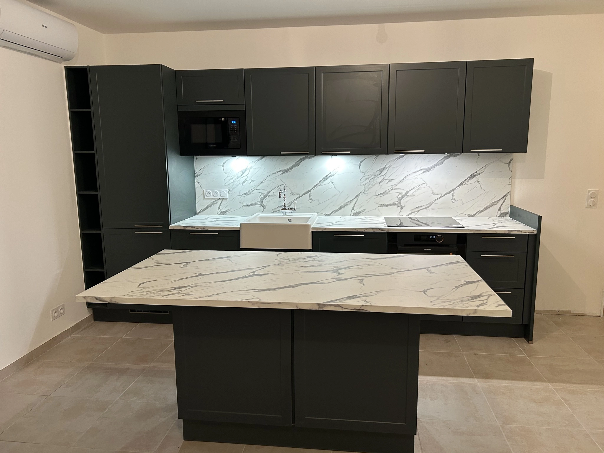 Vue de face d'une cuisine équipée Venidom aux teintes vert foncé (black green) et blanc marbré. L'agencement comprend un linéaire complet avec électroménager encastré et un îlot central spacieux servant de zone de préparation et de partage. Le sol est en carrelage beige clair.