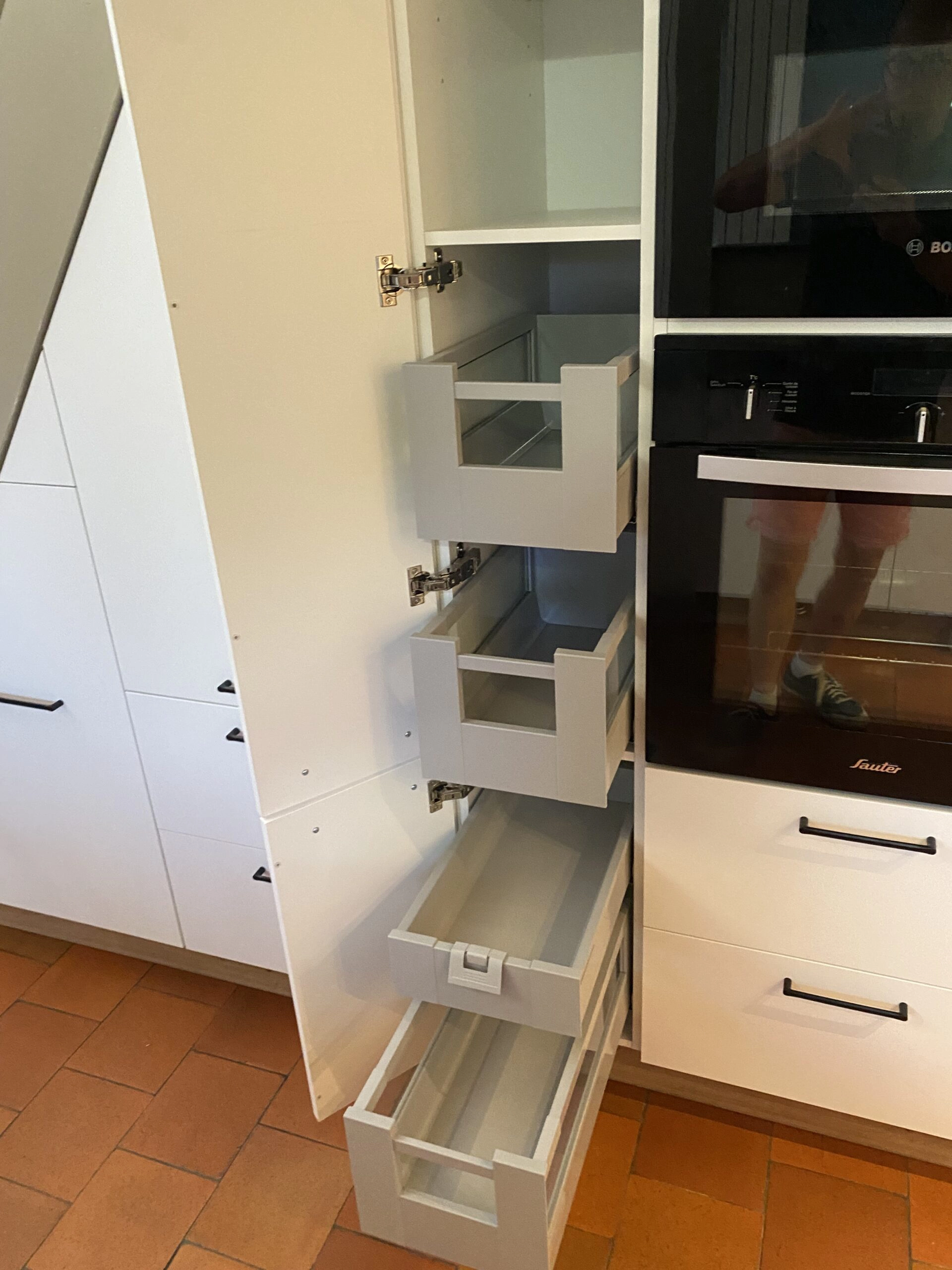 Vue rapprochée d'une cuisine sur-mesure sous un escalier par Venidom. Un placard haut est ouvert, révélant deux tiroirs coulissants gris clair au design moderne. À droite, un four noir de marque Bosch est encastré. Le sol est en carrelage de terre cuite orange.