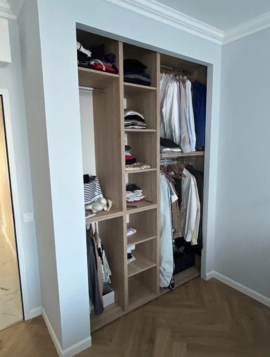 Photo "Avant" d'un dressing ouvert dans une chambre. On y voit des étagères et des penderies en bois remplies de vêtements, créant un désordre visuel. Le sol est un parquet en chevrons et les murs sont bleu clair.