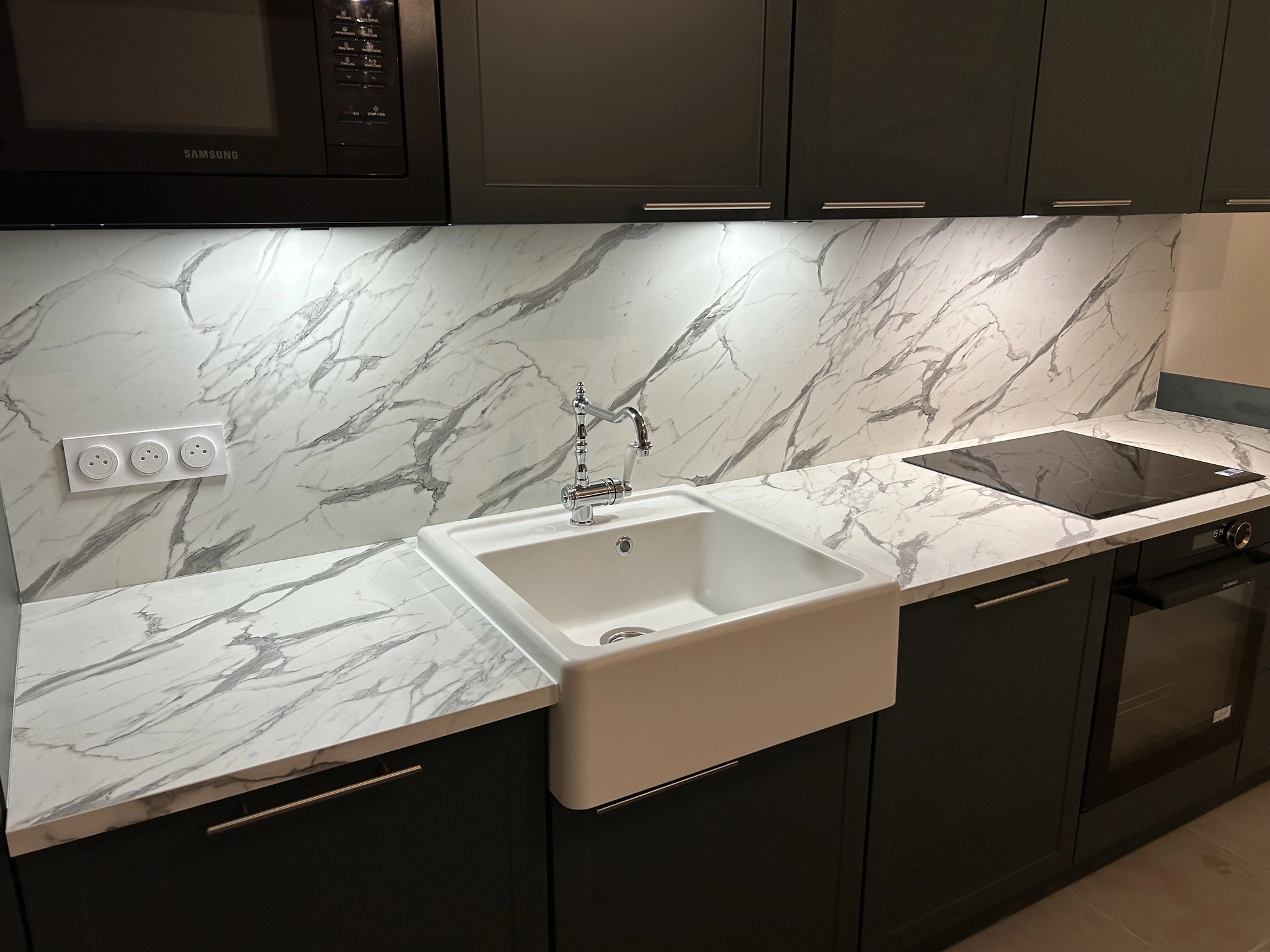 Détail d'une cuisine moderne avec façades mates coloris black green et plan de travail stratifié effet marbre blanc. Un évier timbre blanc traditionnel avec mitigeur rétro chromé apporte du caractère. La crédence assortie au plan de travail est éclairée par des LED sous les meubles hauts.