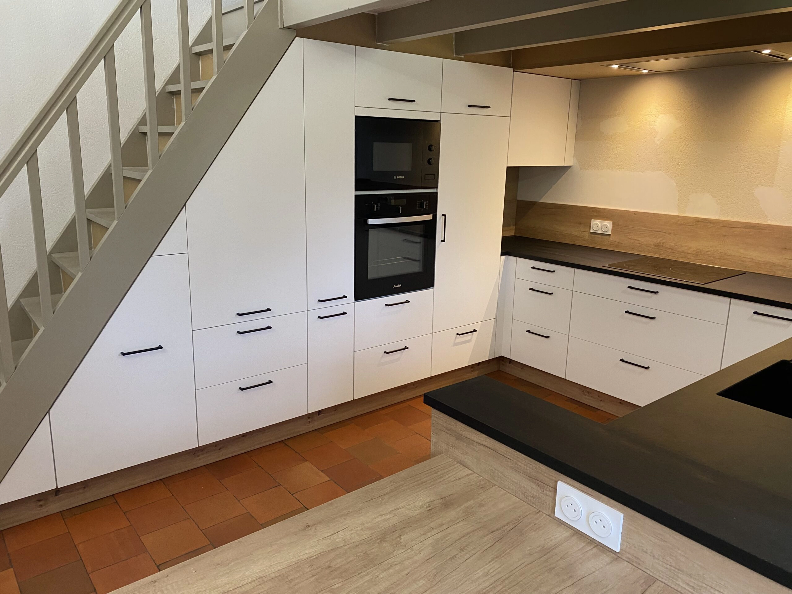 Vue large d'une cuisine en U installée sous un escalier. Elle montre l'optimisation maximale de l'espace avec des rangements sur-mesure blancs sous la pente. Le plan de travail est noir mat avec une plaque de cuisson induction et un évier encastré. Au premier plan, un îlot central avec un coin snack effet bois chaleureux et une double prise de courant blanche. Un projet Venidom Grand Littoral.