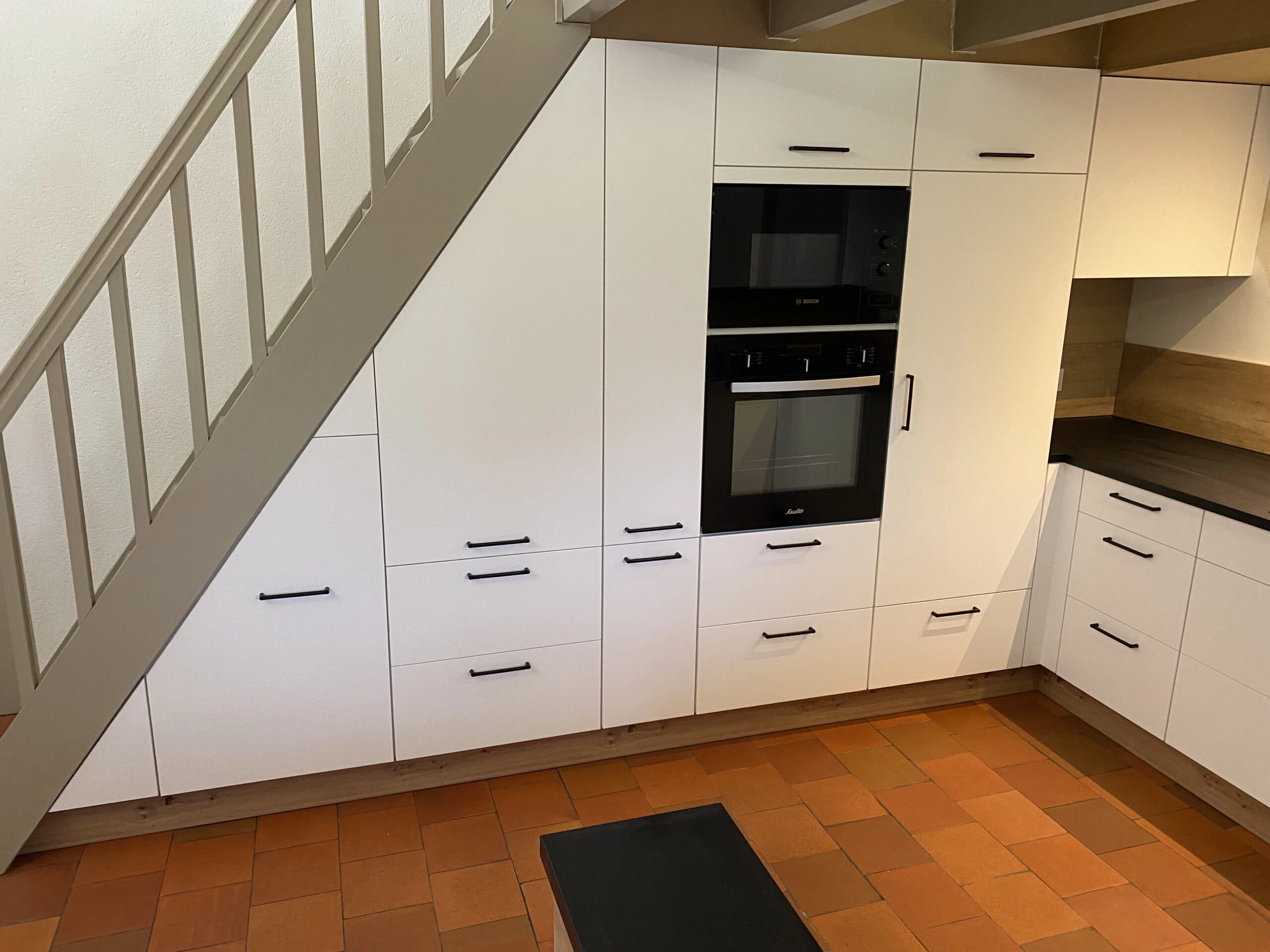 Vue d'ensemble d'une cuisine XXL Venidom entièrement intégrée sous la pente d'un escalier gris clair. Les façades sont blanches épurées avec des poignées noires minimalistes. On distingue les colonnes de rangements, le four et le micro-ondes encastrés, ainsi que le retour en L avec le plan de travail noir. Le sol est en tomettes orange.