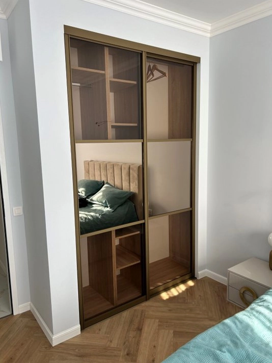 Photo "Après" montrant le dressing structuré par deux grandes portes coulissantes. Les portes possèdent des panneaux en verre bronze et un miroir central reflétant le lit. Les profilés sont en laqué doré poudré. Un projet d'optimisation signé Venidom.