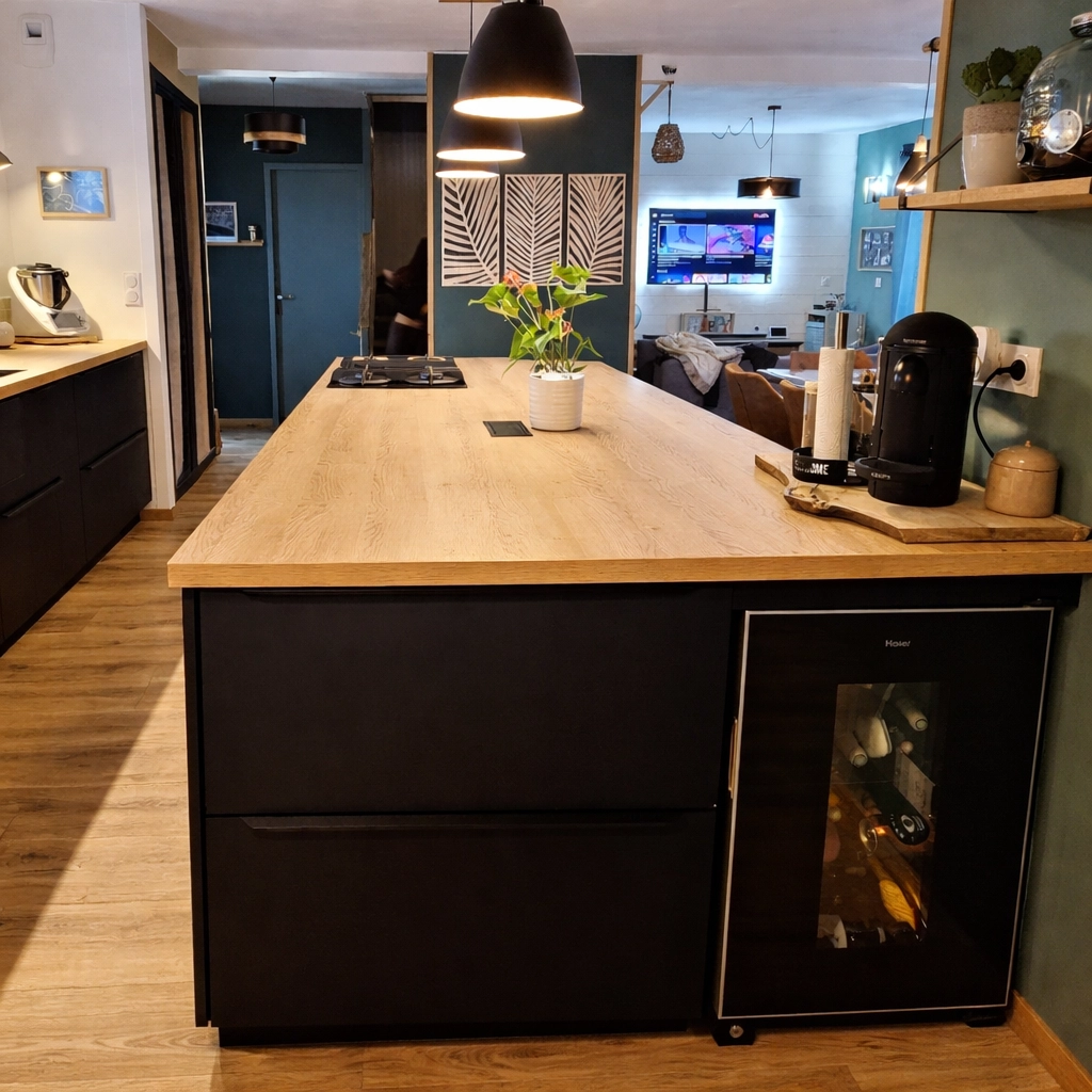 Gros plan sur l'extrémité d'un îlot de cuisine noir mat avec une cave à vin encastrée de la marque Haier. Le plan de travail en bois clair apporte de la chaleur. En arrière-plan, on aperçoit le salon ouvert, créant une transition fluide entre l'espace préparation et l'espace de vie.