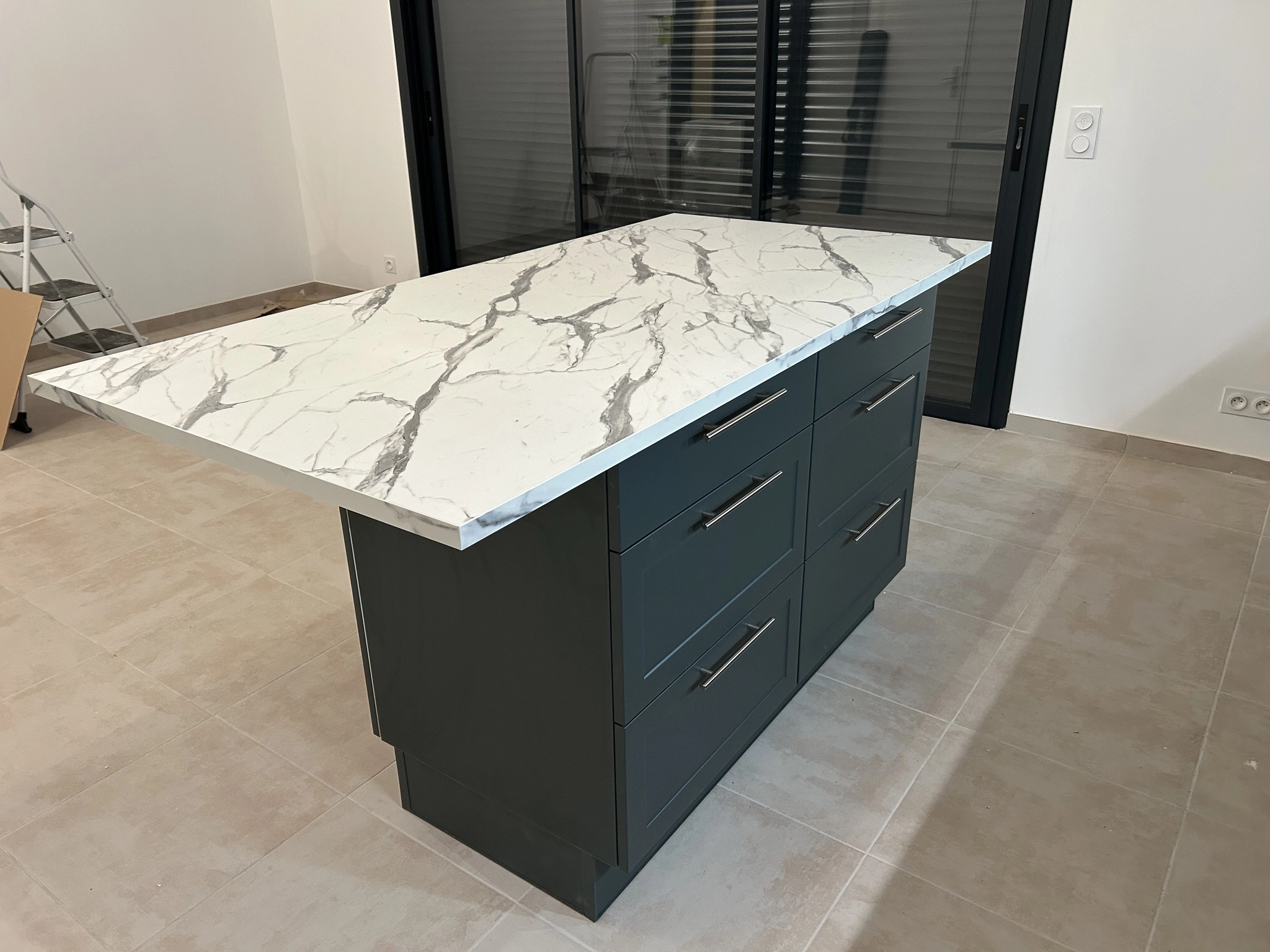 Gros plan sur l'îlot central d'une cuisine contemporaine. Il dispose de larges tiroirs de rangement avec poignées fines en inox sur des façades vert forêt mat. Le plan de travail débordant en effet marbre blanc permet de créer un coin repas. En arrière-plan, une baie vitrée avec volets roulants noirs.