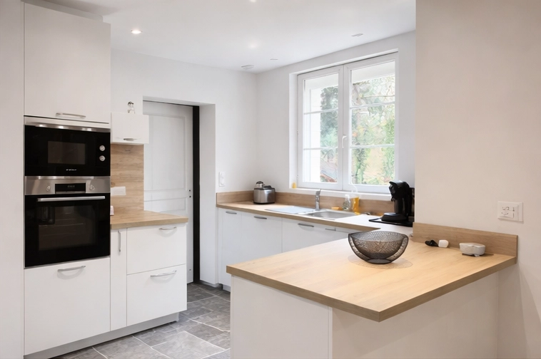 Aménagement d'une petite cuisine lumineuse par Venidom avec des façades blanches épurées et un plan de travail en bois clair. Au premier plan, un îlot convivial avec un bol design. En arrière-plan, l'évier est placé sous une fenêtre offrant une vue sur l'extérieur. Le sol est en carrelage gris effet pierre.