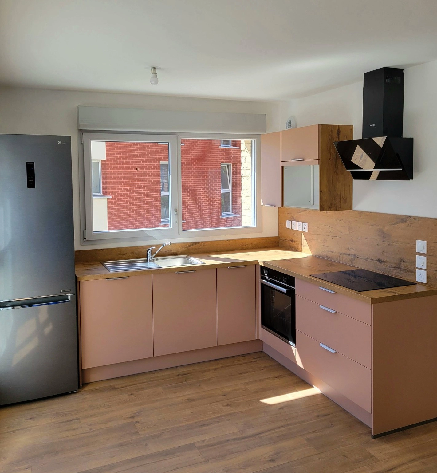 Petite cuisine moderne en L avec façades dynamiques rose orangé, électroménager intégré et crédence effet bois, conçue par Maximilien Lust (Venidom Grand Littoral).
