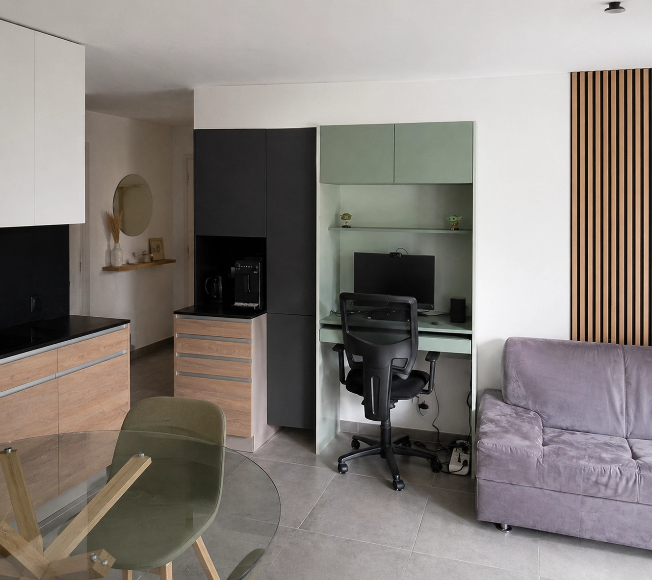 Vue d'ensemble montrant l'intégration parfaite d'un bureau vert sauge entre une cuisine moderne noire et bois et un salon avec canapé lilas et panneau décoratif en tasseaux de bois.