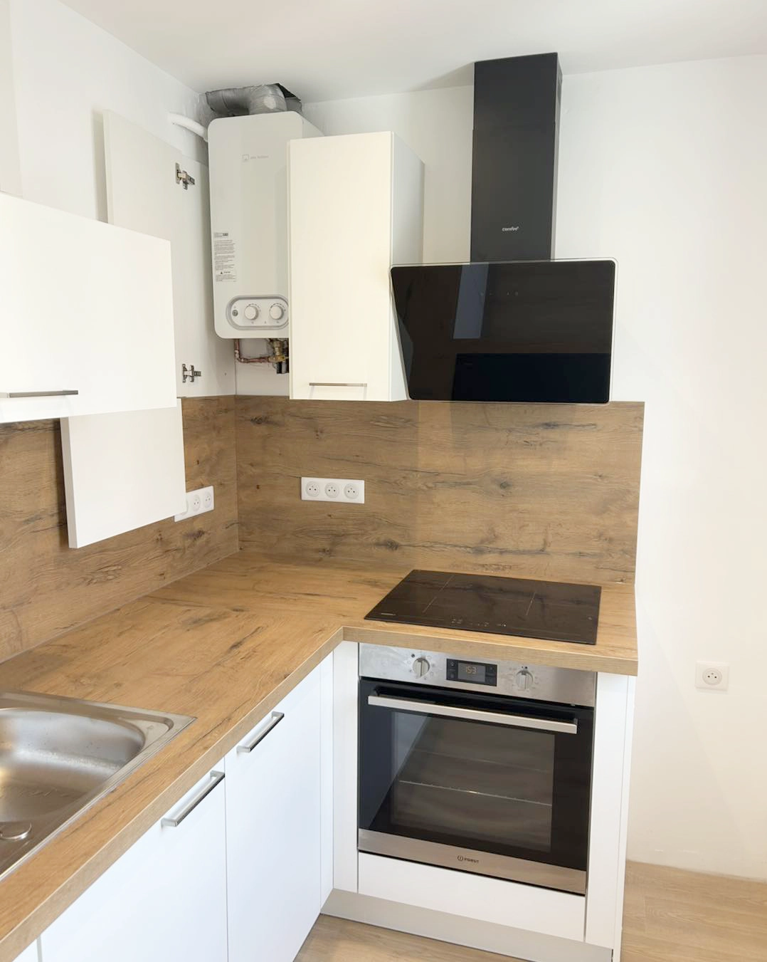 Cuisine compacte en angle avec façades blanches et crédence bois, rangements optimisés et électroménager intégré dans un petit espace