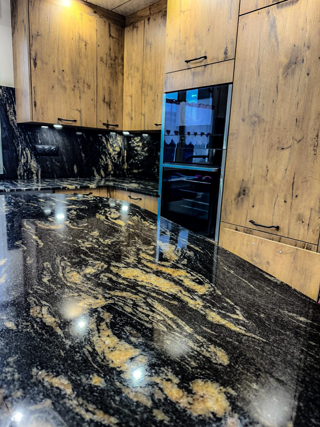 Gros plan sur un îlot central au plateau en granit noir brillant avec des veines or et ocre complexes. En arrière-plan, une colonne d'armoires en bois de style rustique intègre un double four encastré en verre noir moderne et un éclairage LED intégré sous les meubles hauts. Un projet signé Venidom Grand Littoral.