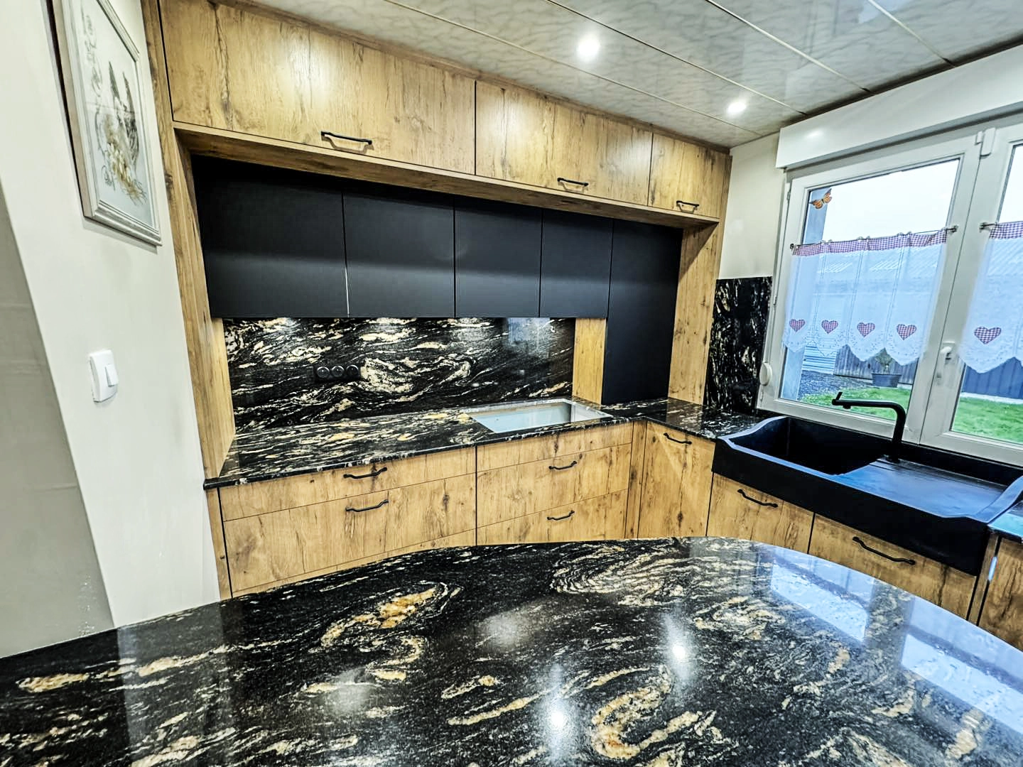 Gros plan sur un plan de travail en granit noir brillant veiné de doré, avec une plaque de cuisson à induction encastrée et un évier encastré en inox. Le contraste est fort entre la crédence en marbre foncé, les meubles bas effet bois et des rangements noirs mats épurés au centre. Le sol en carrelage beige est visible.