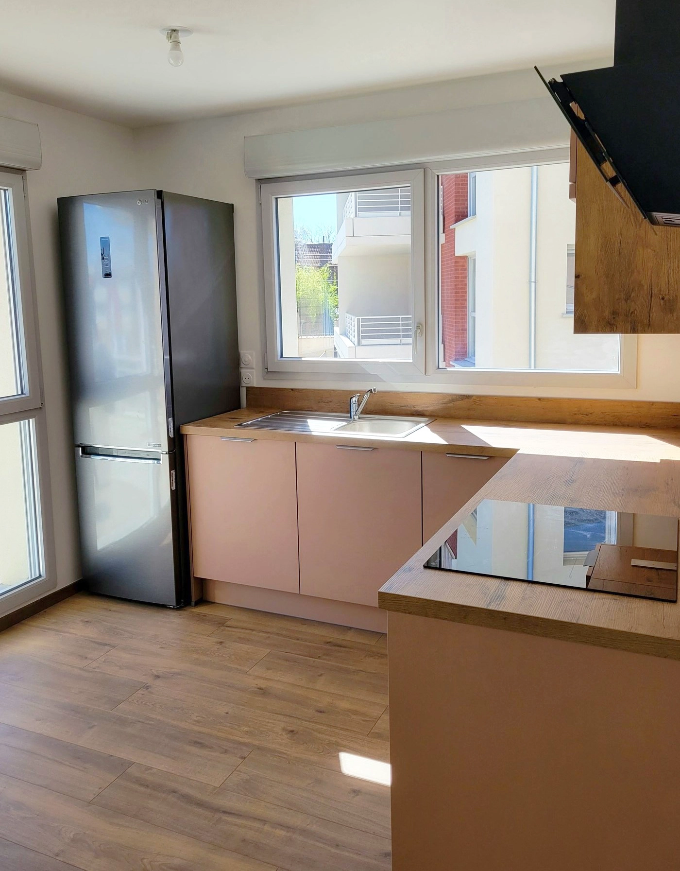 Aménagement d'une petite cuisine baignée de lumière avec façades rose corail, plan de travail effet bois et évier inox situé sous une fenêtre.