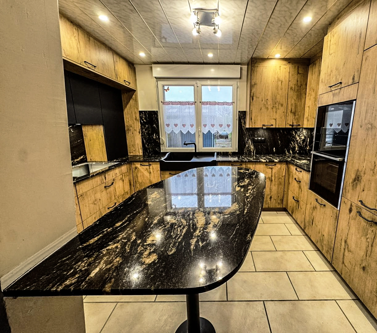 Vue d'ensemble d'une cuisine moderne rénovée en 5 jours par Venidom. Elle présente un contraste élégant entre des façades effet bois chaleureux et un îlot central, plan de travail et crédence en granit ou marbre veiné or. Un évier noir est placé sous une fenêtre ornée de rideaux à cœurs. Le sol est composé de grands carreaux beiges.