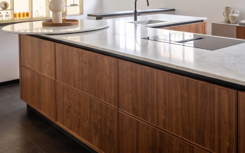 Îlot de cuisine haut de gamme en bois de meuble foncé surmonté d'un plan de travail en marbre blanc veiné avec plaque de cuisson intégrée.