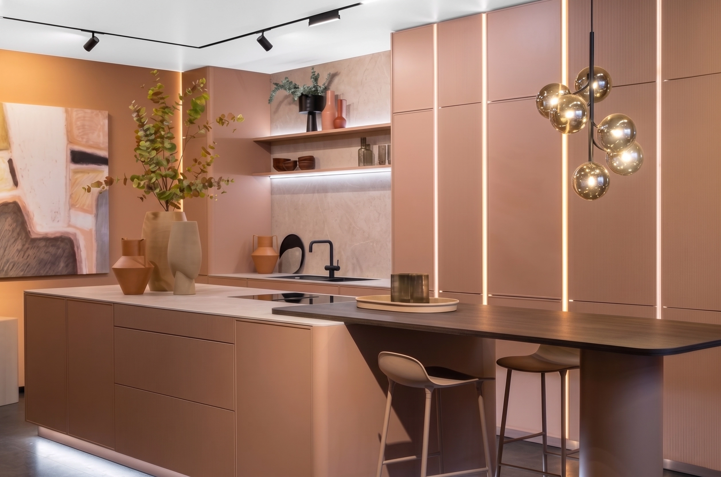 Cuisine moderne haut de gamme aux façades rose poudré avec un grand îlot central, des luminaires suspendus dorés et des rangements rétroéclairés par des LED.