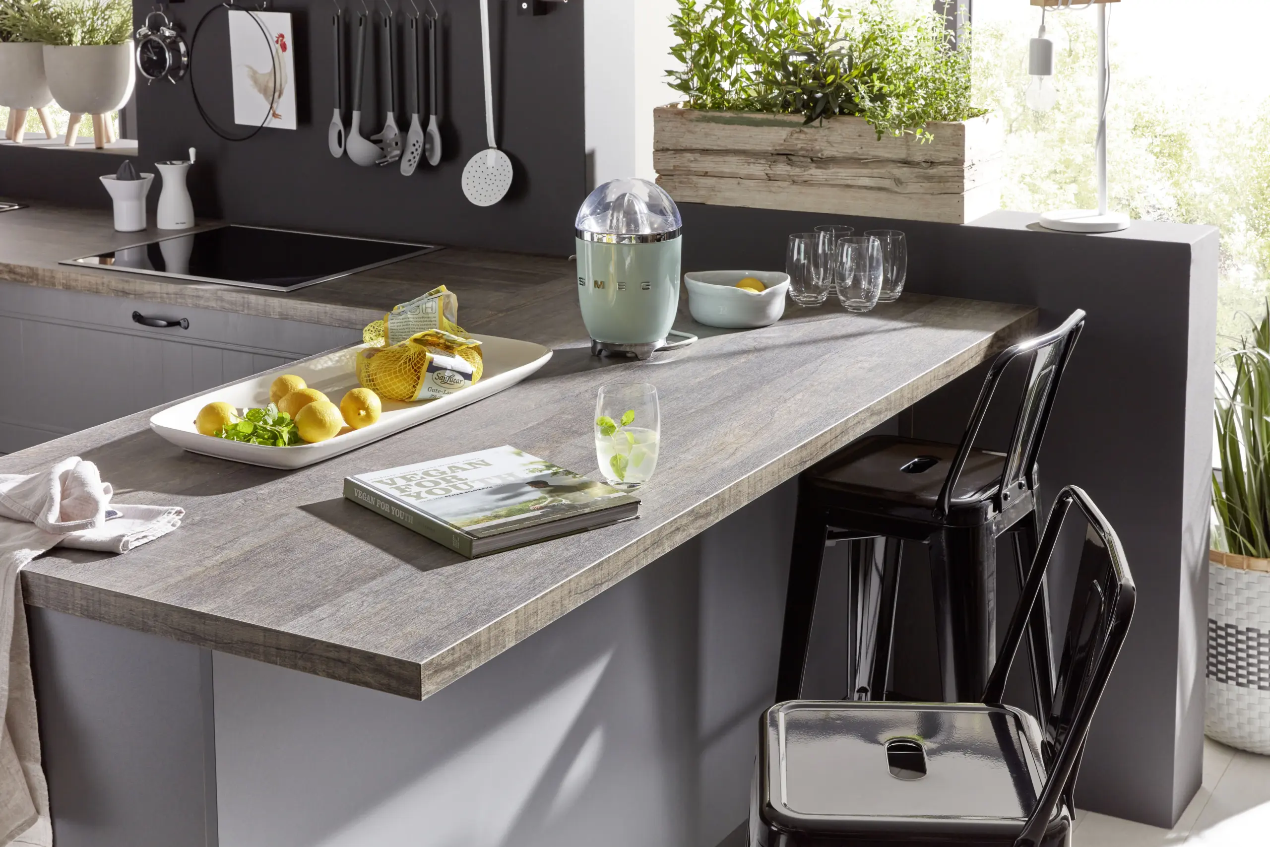 Zoom sur un plan de travail de cuisine effet bois foncé sur un îlot gris, décoré avec des citrons, un livre de cuisine et des tabourets industriels noirs.